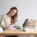 Dievča sedí pri stole s notebookom a knihami. Má online doučovanie a píše si do zošita poznámky.